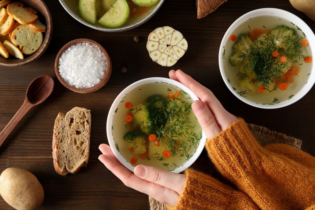 A mug of clear broth held in two hands on an empty table, the concentrated form that raises blood glutamate fastest.
